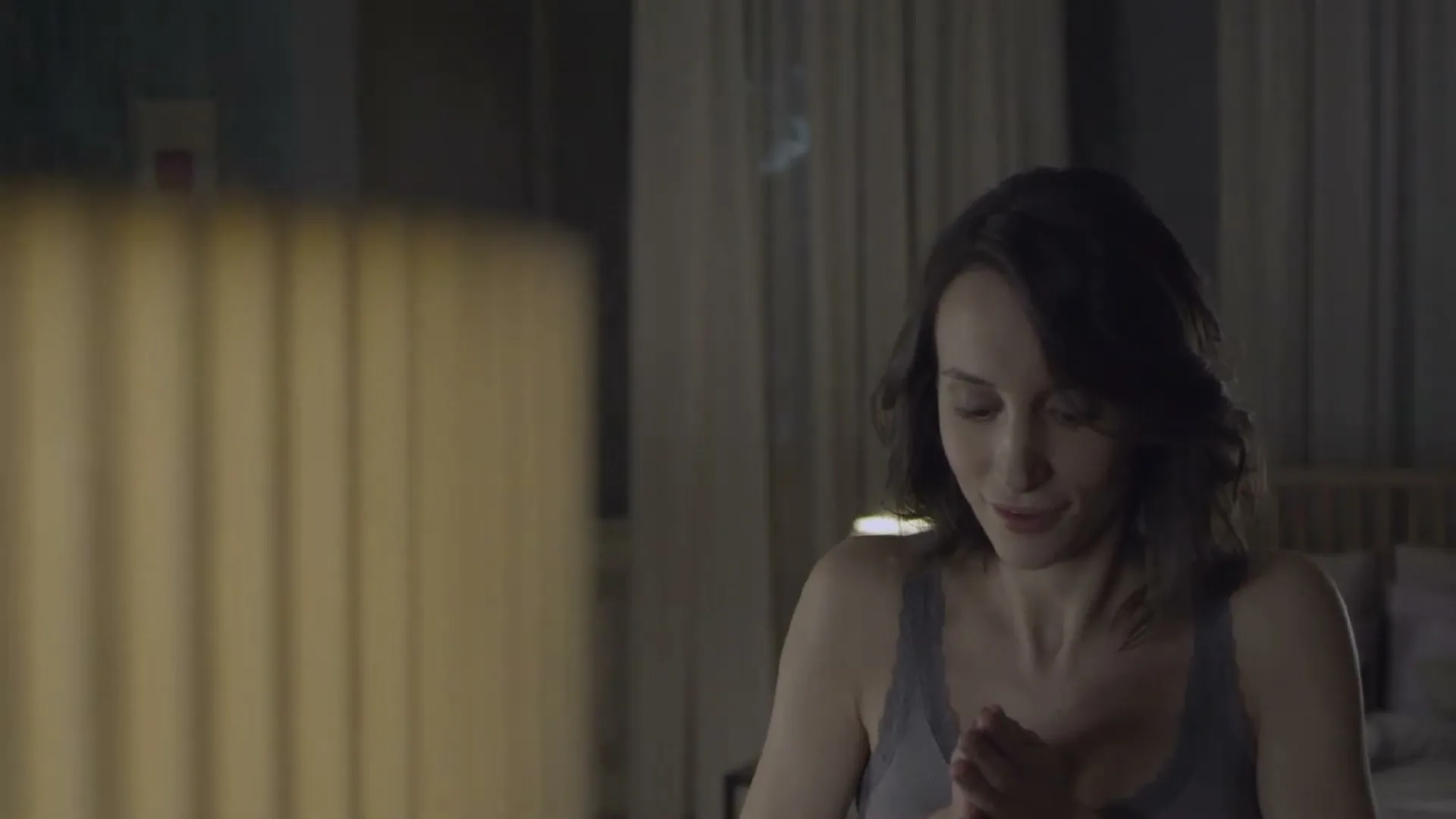 A woman in her 30s, wearing a grey tank top, looks surprised and then relieved in a dimly lit bedroom.