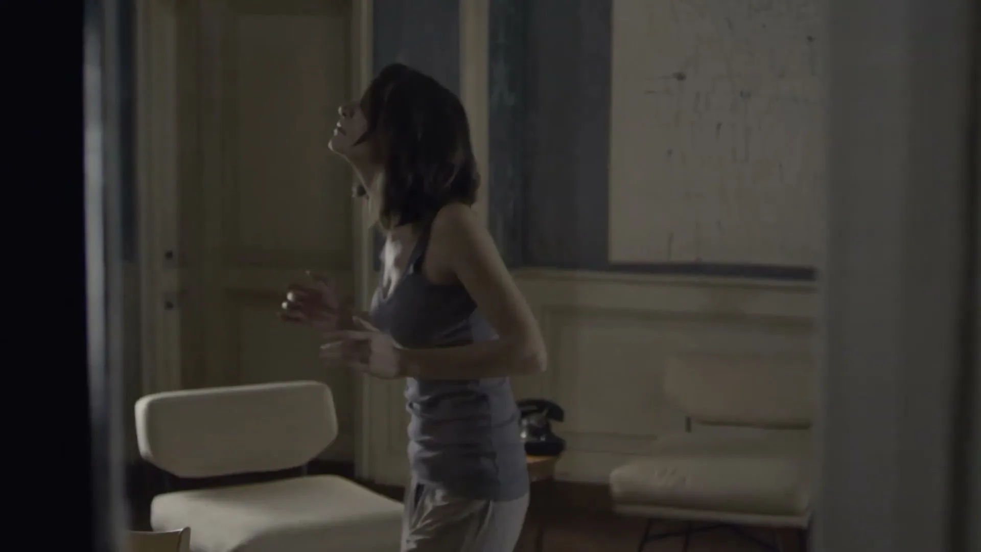 A young woman in a tank top and shorts stands in a dimly lit room, looking up and moving her hands as if applying something to her face.