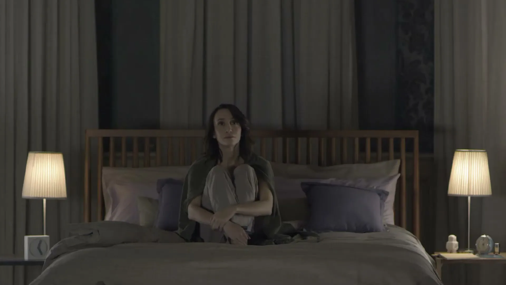 A woman sits alone on a bed in a dimly lit bedroom, looking thoughtful.