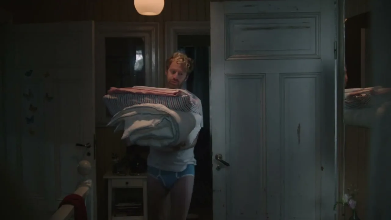 A man with curly blonde hair, wearing a white t-shirt and blue briefs, walks through a dimly lit bedroom carrying a stack of bedding.