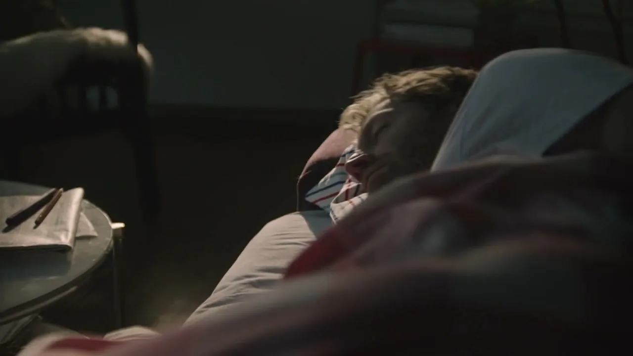 A man with light brown hair and a white t-shirt is sleeping in bed, then wakes up and looks around in a dark bedroom.