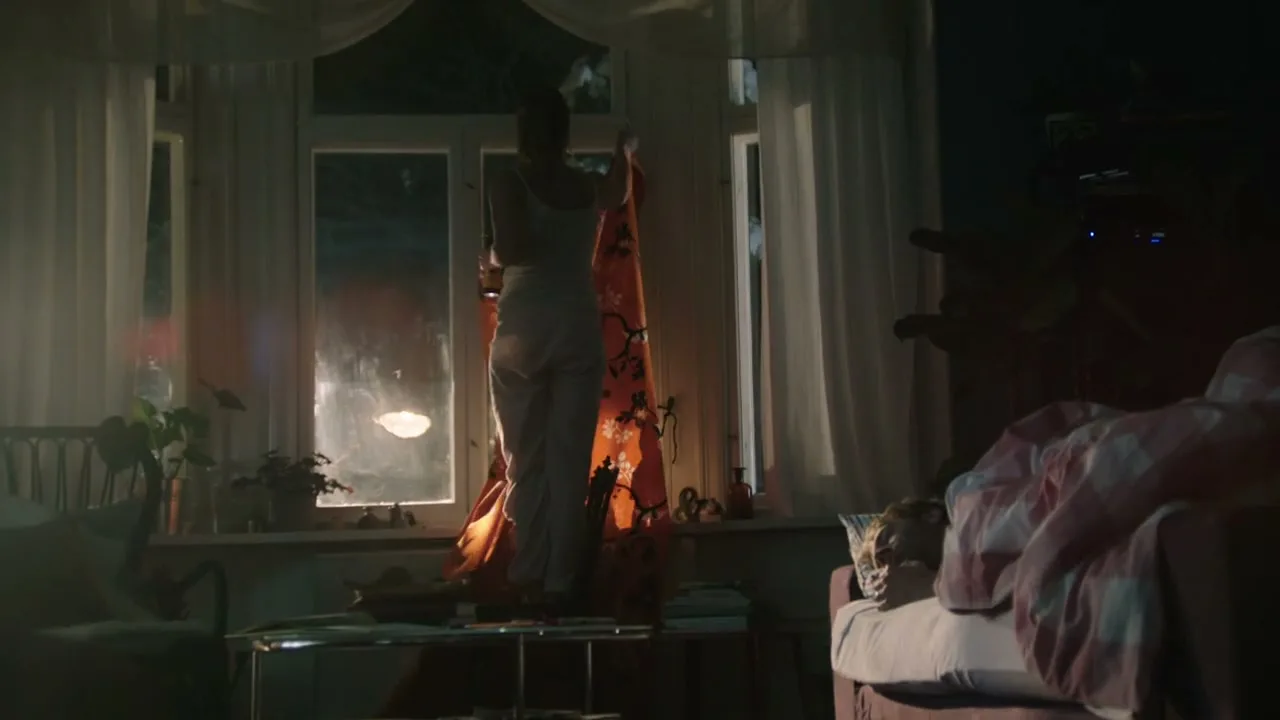 A woman in pajamas stands on a table, holding up a patterned sheet to cover a window, while a man sleeps in bed in a dimly lit bedroom.