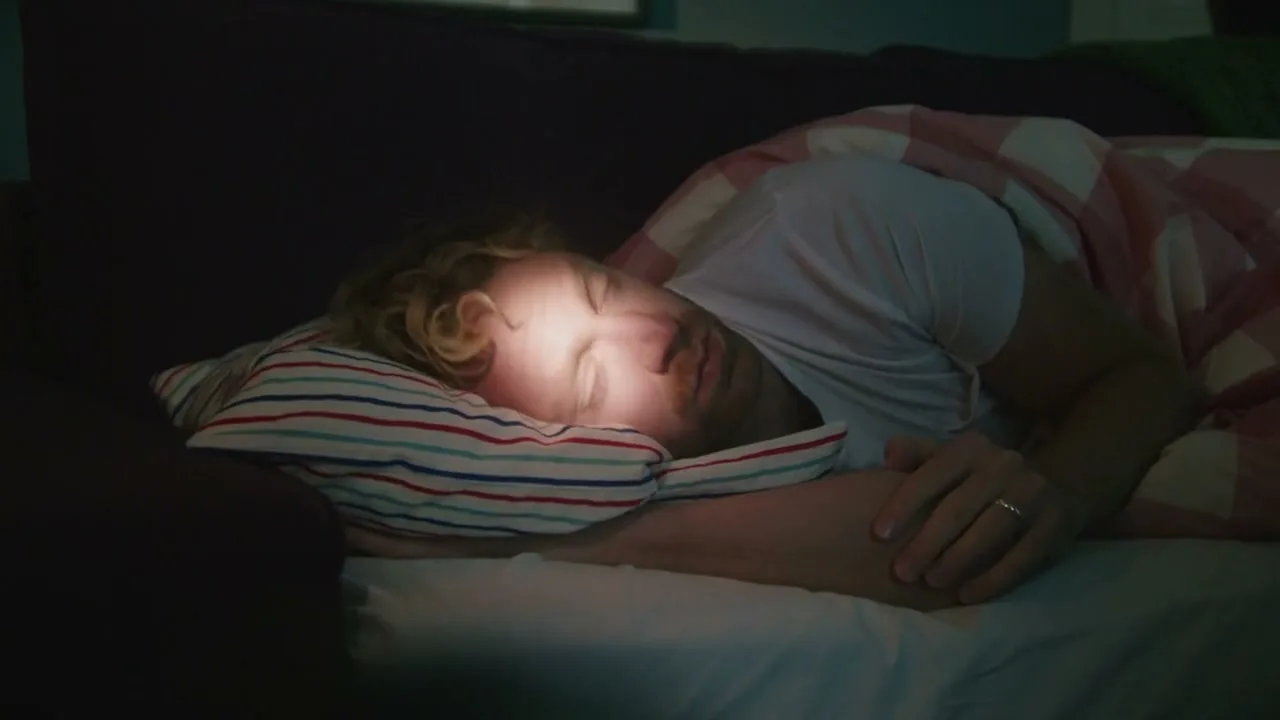 A man with curly blonde hair sleeps soundly in bed, illuminated by a soft light, in a dark bedroom.