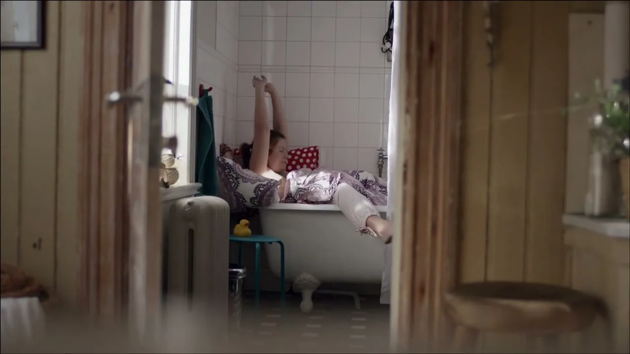 A young woman in pajamas stretches in a bathtub, seen through a doorway in a cozy bathroom during the day.