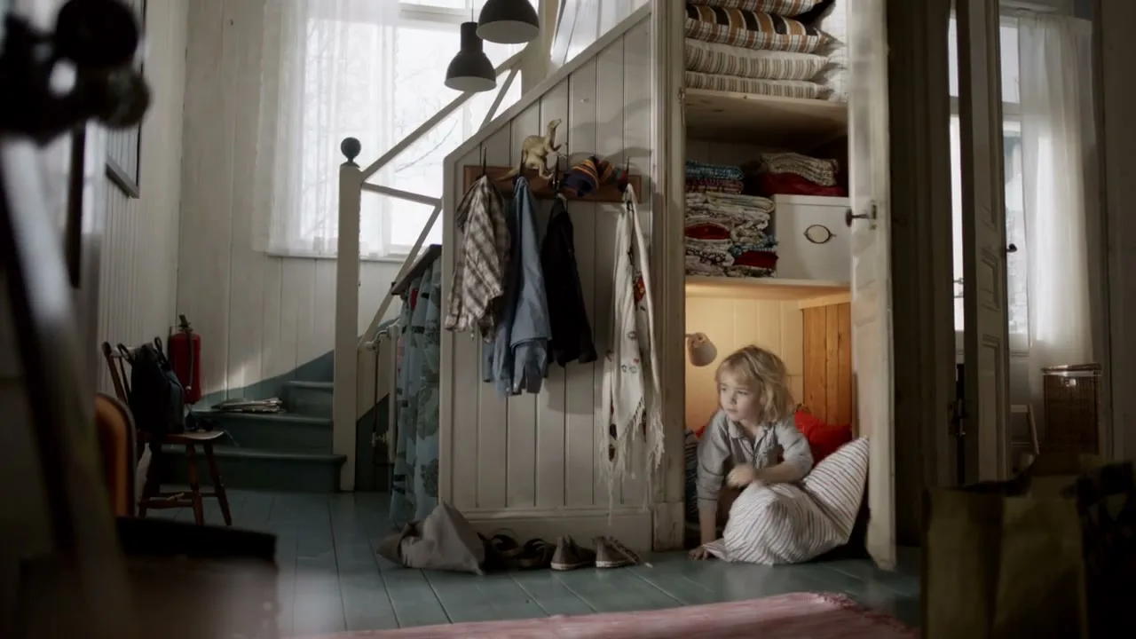 A young girl with blonde hair in pajamas peeks out from a closet filled with pillows and blankets in a cozy, sunlit hallway.