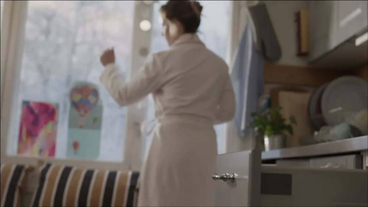 A cat jumps off a woman's shoulder onto a kitchen drawer, then the woman in a bathrobe looks out a window in a bright kitchen.