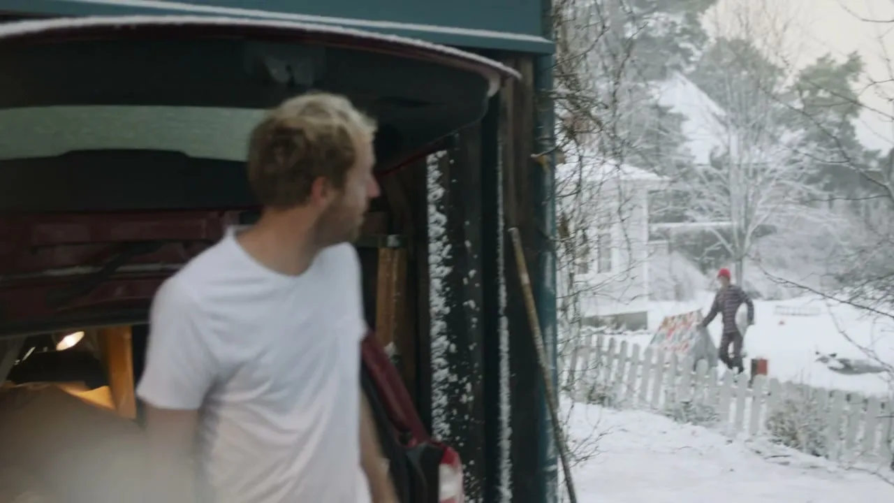 A man in his underwear and t-shirt gets out of the back of a car in a snowy backyard and waves to another man.
