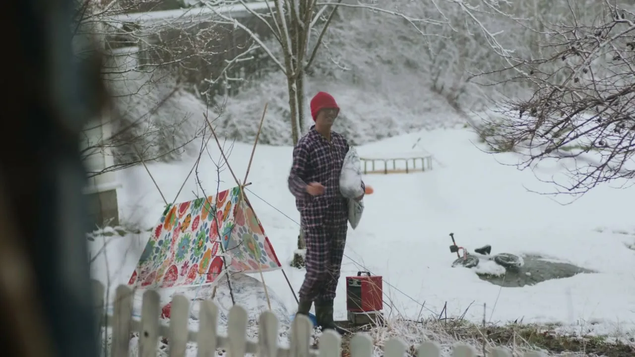 A man in red pajamas and a red hat waves goodbye while holding a pillow, standing next to a colorful tent in a snowy backyard.