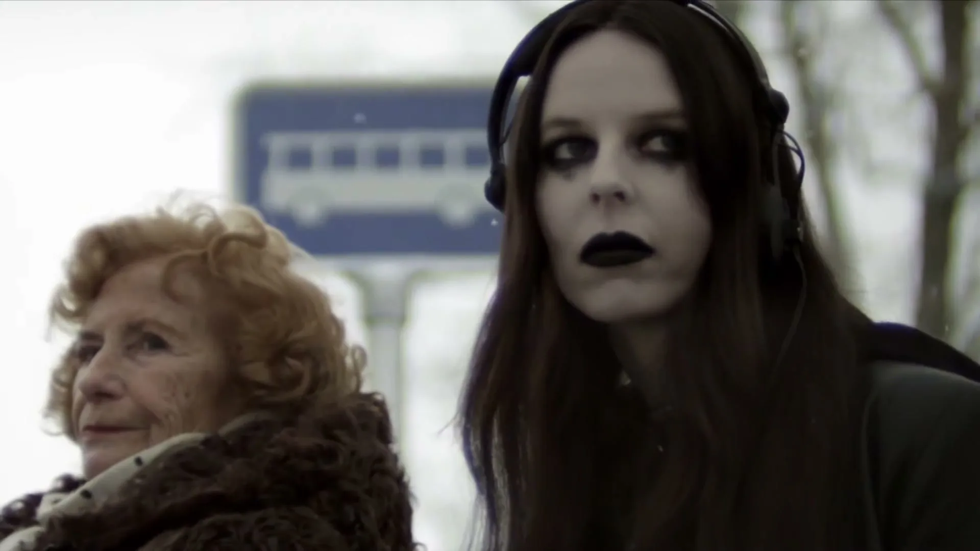 An elderly woman with curly red hair and a young woman with long dark hair, pale skin, and dark makeup wait at a bus stop on a snowy day.