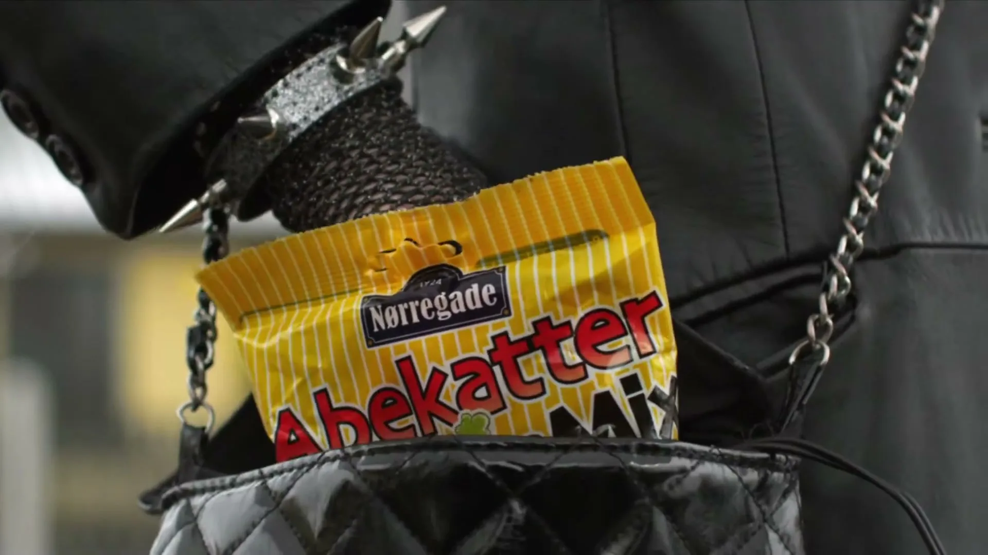 A pale hand with a ring and fishnet glove pulls a yellow bag of Nørregade Abekatter Mix candy out of a black quilted purse.