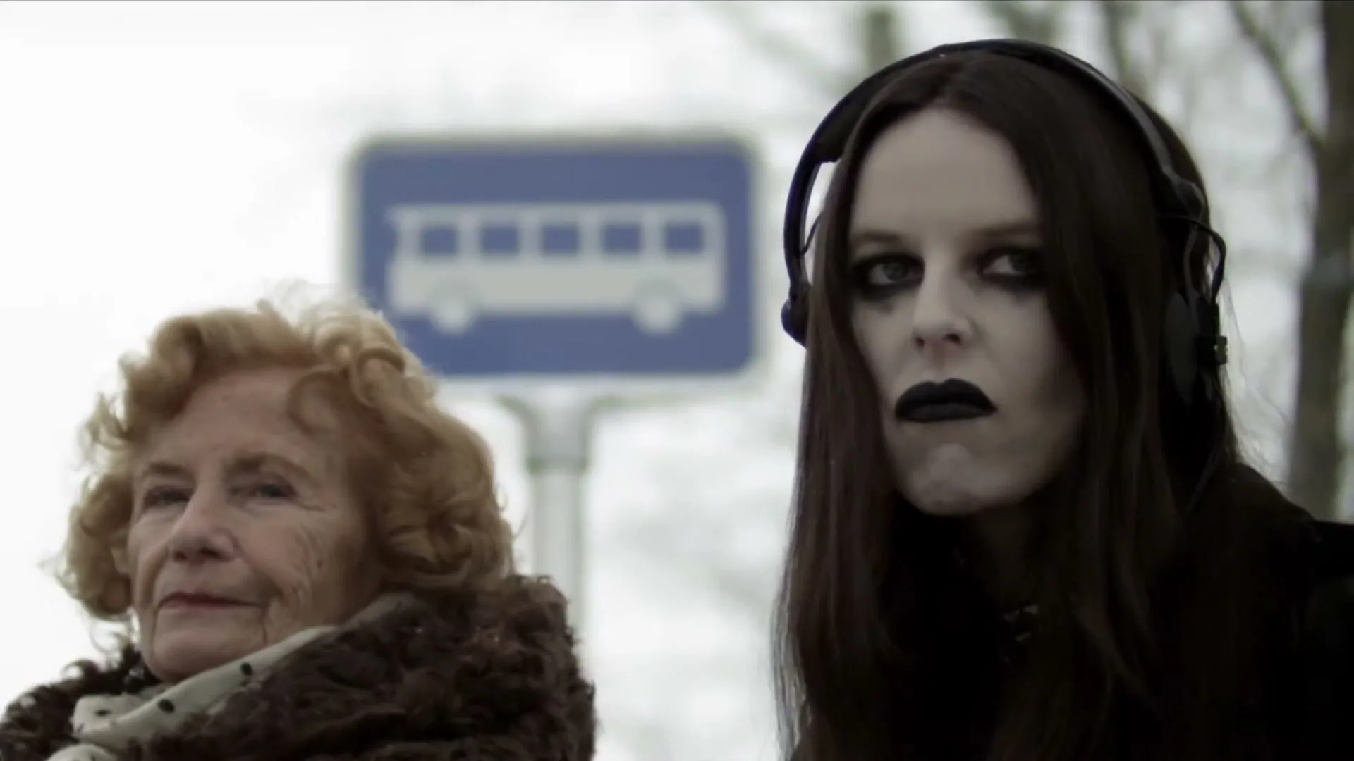 A goth woman with headphones and an elderly woman sit at a bus stop, with the goth woman eating candy and making a face.