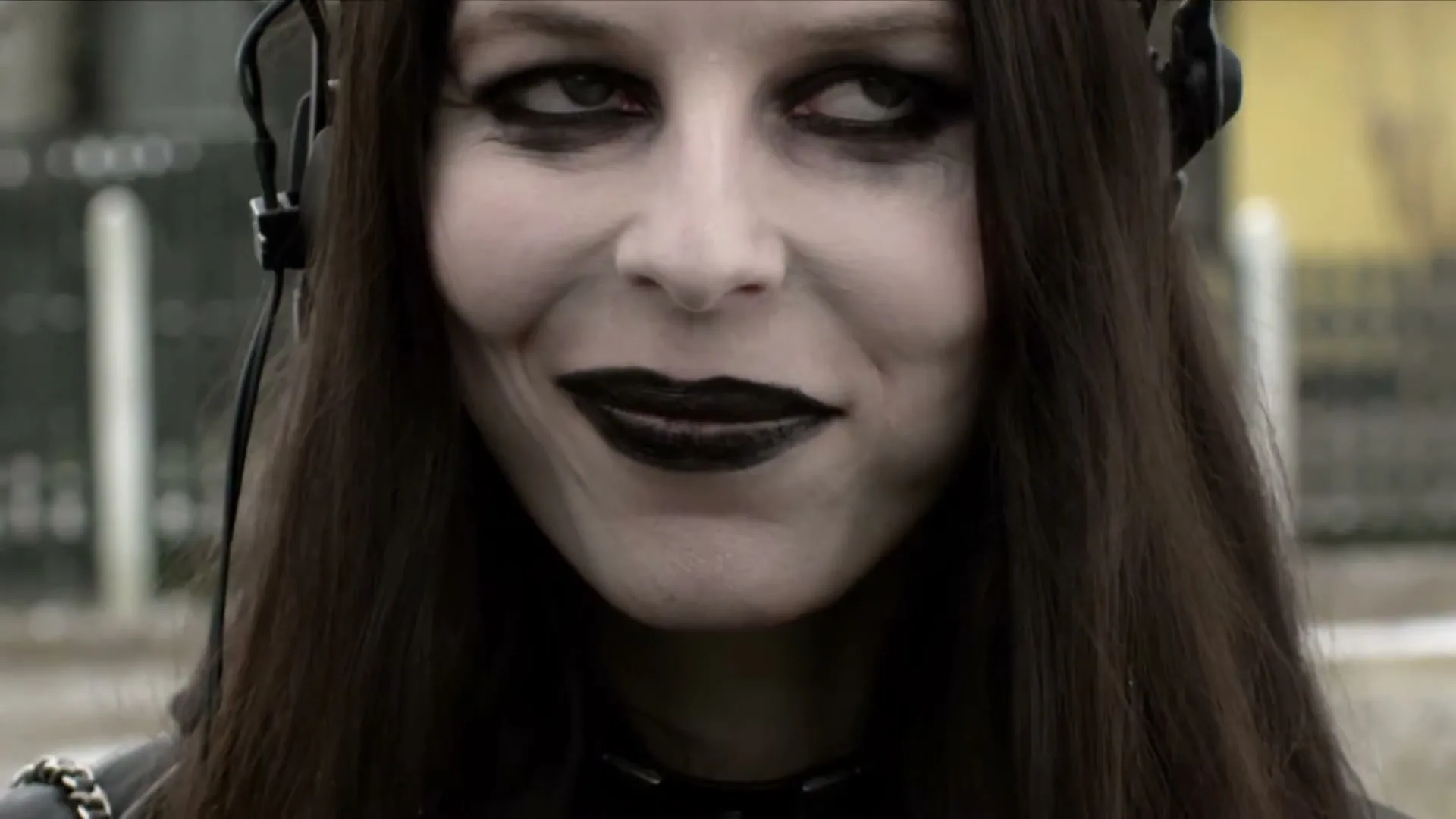 A pale Goth woman with dark makeup and headphones smiles subtly, then broadly, as she listens to something outdoors.