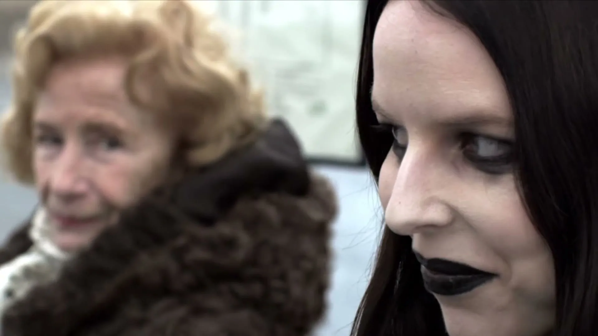 A pale woman with dark makeup and hair looks at an older woman with curly blonde hair and a fur coat, who is looking back at her, at a bus stop.