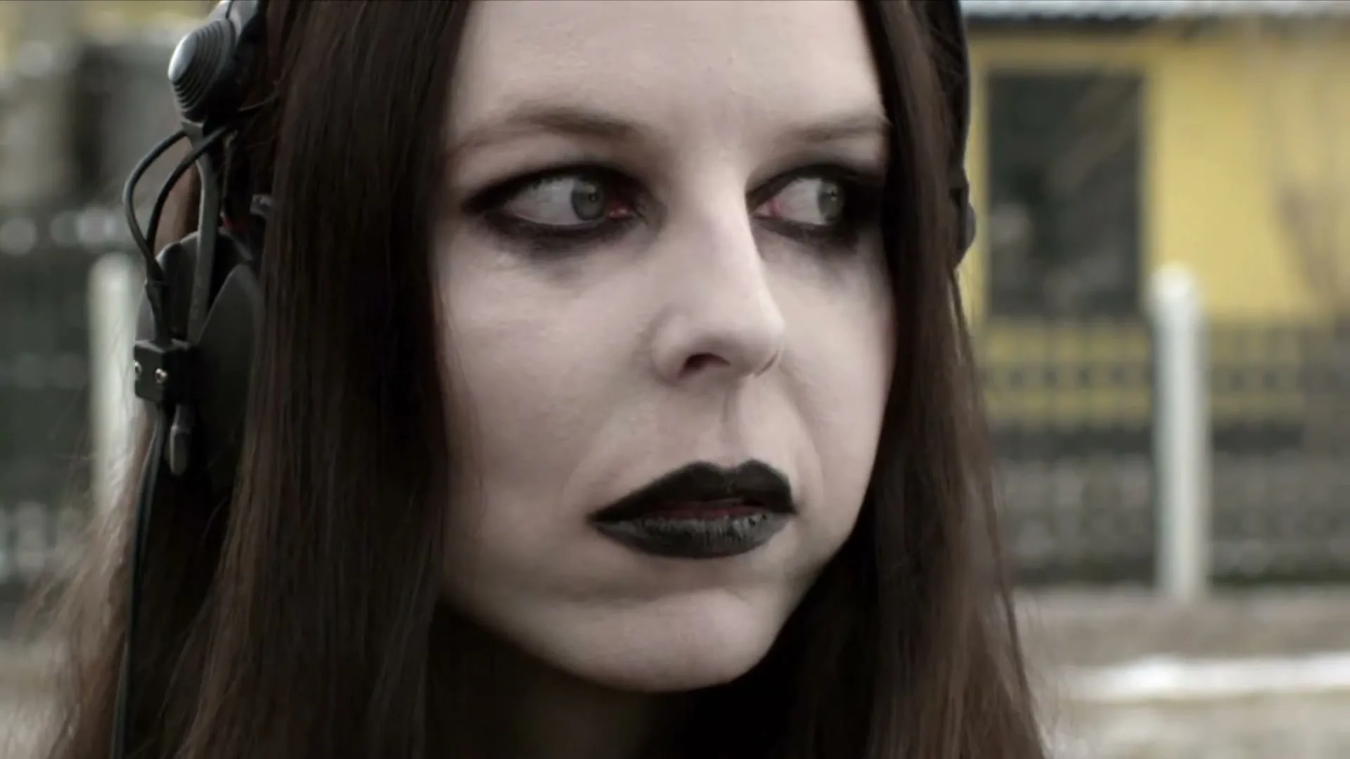 A young woman with pale skin, dark hair, and black lipstick, wearing headphones, looks around nervously in an outdoor setting.