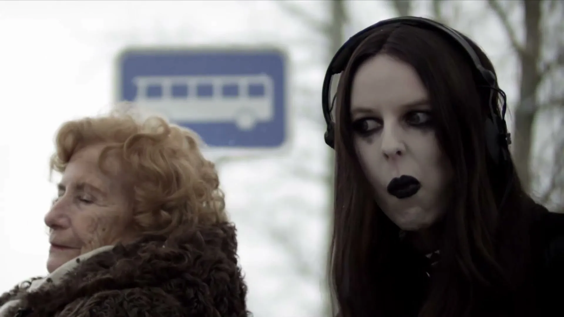 A goth woman with headphones and an older woman in a fur coat wait at a bus stop in a snowy, outdoor setting.