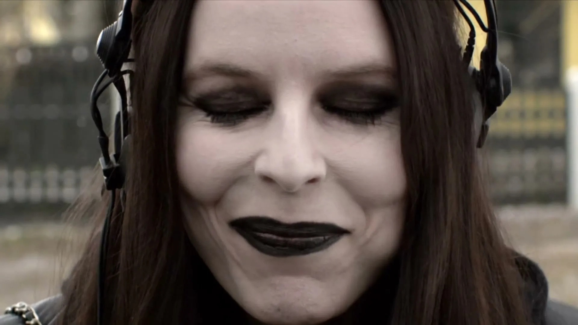 A pale woman with dark hair and gothic makeup, wearing headphones, closes her eyes and smiles, then opens them and looks around, outdoors.