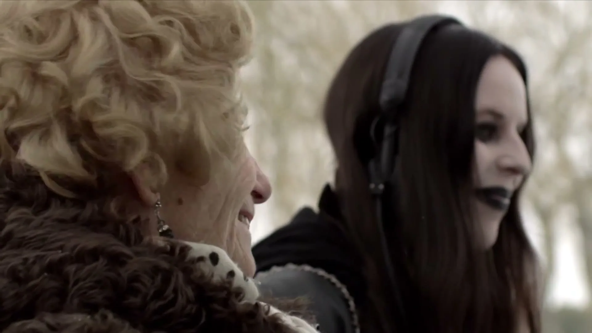 An older woman with curly blonde hair and a younger woman with long dark hair and headphones are seen from behind, both smiling slightly outdoors.