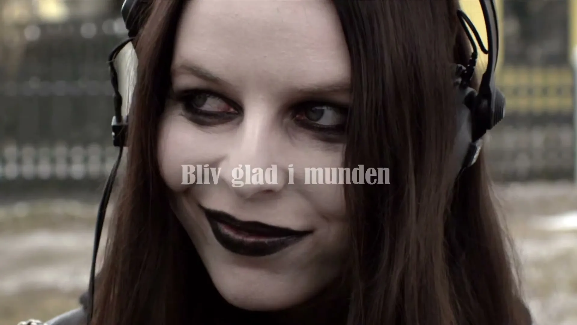 A pale woman with dark hair and makeup, wearing headphones, smiles slyly while looking to the side, with text overlay.