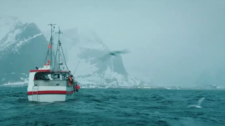 A fishing boat with two people on board navigates choppy waters with snow-capped mountains in the background on a cloudy day.