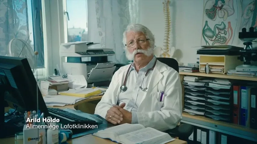 An older male doctor with a white mustache and glasses sits at a cluttered desk in his office, speaking directly to the camera.