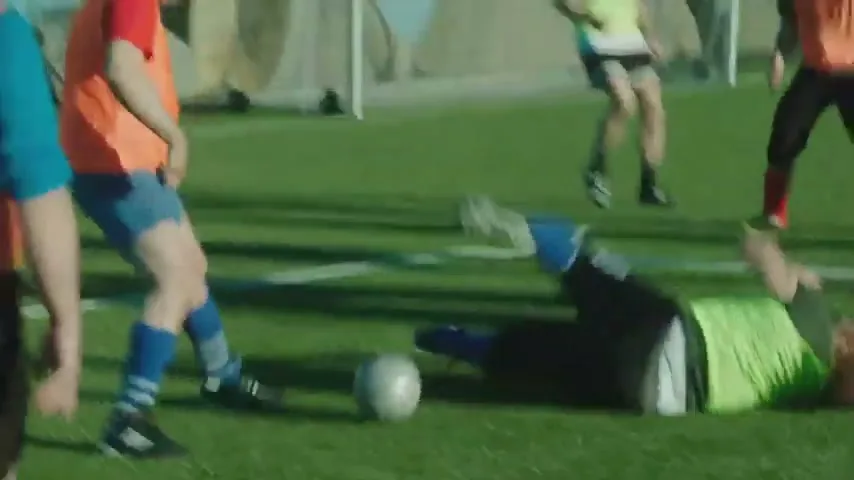 A group of adults play soccer on a green field with mountains in the background, and one player falls to the ground in pain.