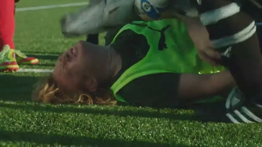 A male soccer player in a neon green vest lies on the ground in pain, clutching his knee, while other players' legs are visible around him on a sunny day.
