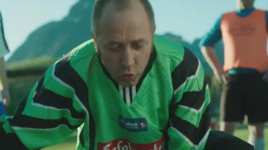 A male soccer player in a green jersey is hunched over, looking down, on a sunny outdoor field.