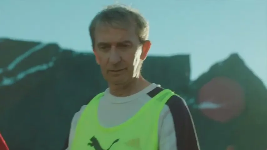 A middle-aged man in a neon green bib stands on a sunny outdoor soccer field, looking down with a serious expression.