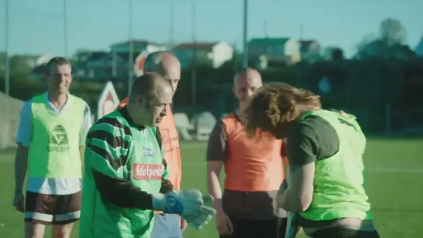 A group of middle-aged men in soccer jerseys and vests gather on a sunny green field, with one man in a green goalie shirt looking down at something in his hands.