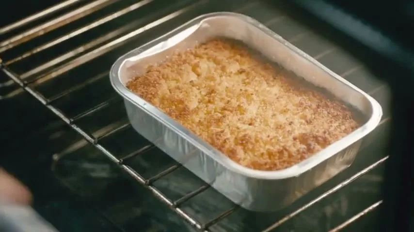 A close-up shot shows a golden-brown fish gratin in a foil tray, baking inside a dark oven, as a hand in an oven mitt reaches in to remove it.