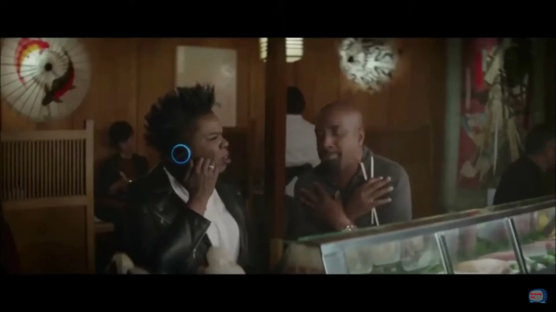 Leslie Jones, a Black woman, wears a black leather jacket and white shirt, talking into a blue glowing device on her ear, while a Black man sits next to her in a sushi restaurant.