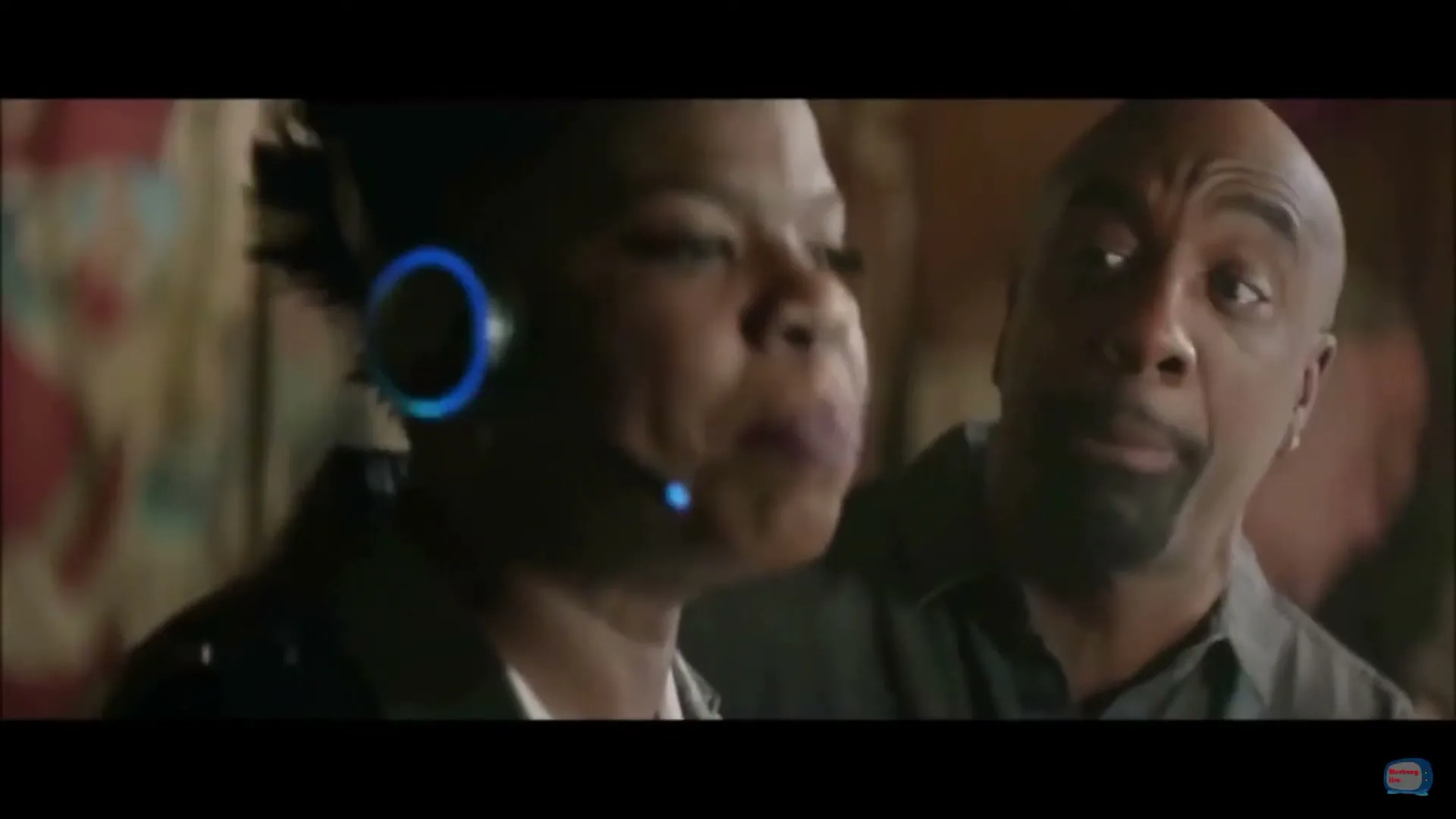 A woman with a headset and a man with a beard are talking animatedly, looking at each other in a dimly lit interior.