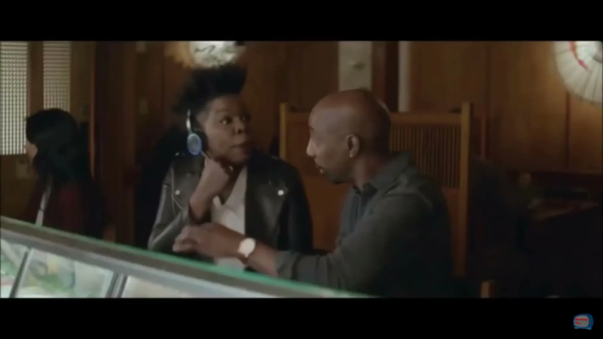 A man and a woman, both in their 30s, sit at a counter in a restaurant, talking to each other.