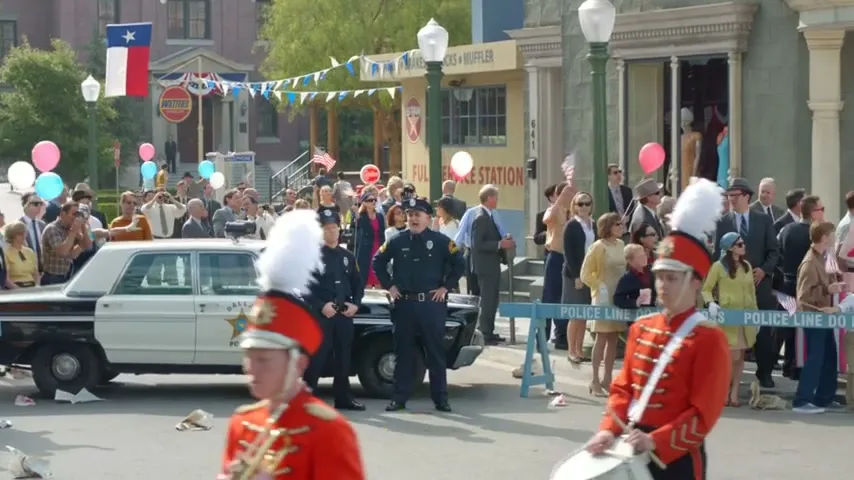 Two police officers stand in front of a police car, watching a parade with a marching band and a crowd of people on a sunny day in a small town.