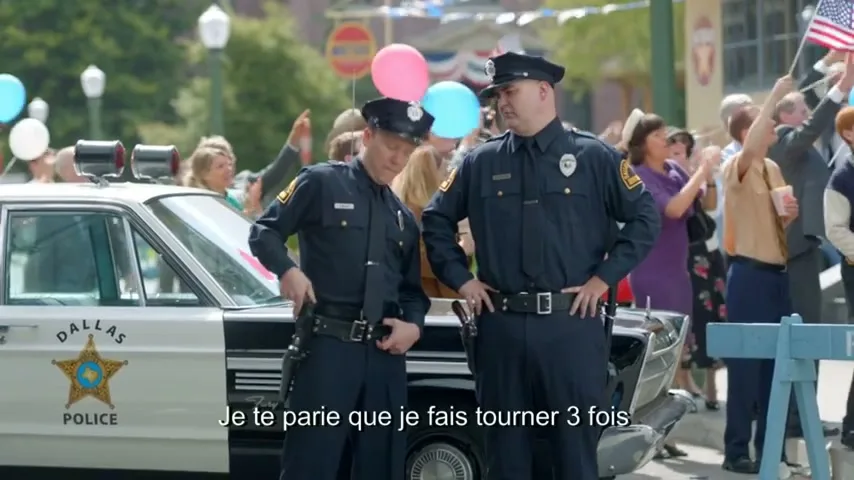 Two police officers in uniform stand next to a Dallas Police car, talking to each other while a crowd of people watches in the background on a sunny day.