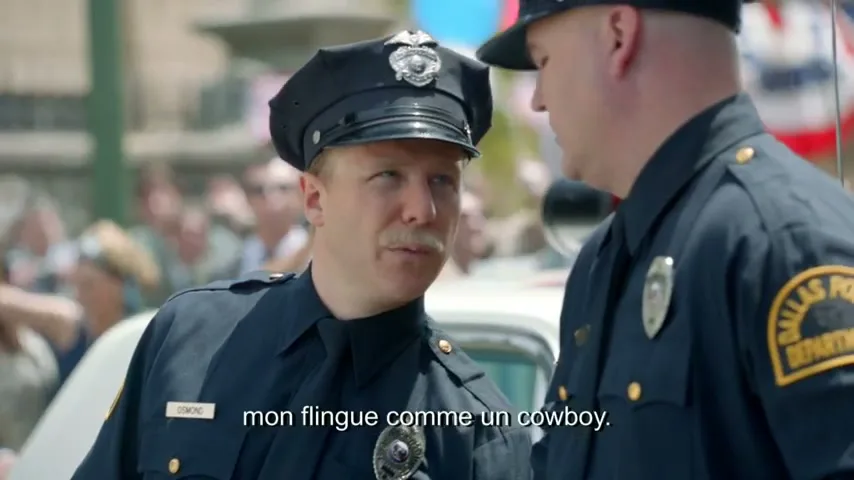 Two male police officers in uniform talk to each other on a sunny day in a street with a crowd in the background.