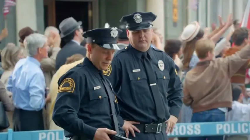 Two male police officers in uniform stand in front of a crowd, one looking down and the other looking forward, in an outdoor setting.