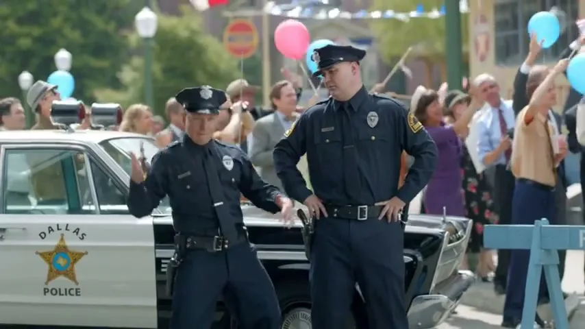 Two male police officers in uniform stand next to a Dallas Police car, one dancing while the other watches, as a crowd celebrates in the background.