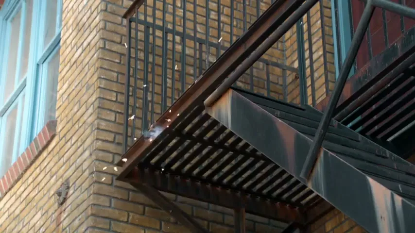 Sparks fly from a metal fire escape attached to a brick building with a blue window, suggesting a struggle or impact.