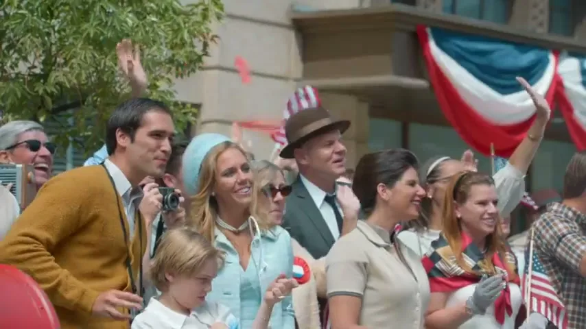 A crowd of people, including a man in a yellow sweater, a woman in a blue top, and a child, wave and smile at a parade on a sunny day.