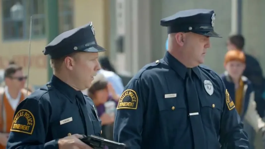 Two male police officers in uniform look around with surprised expressions, one holding a gun, on a busy street during the day.