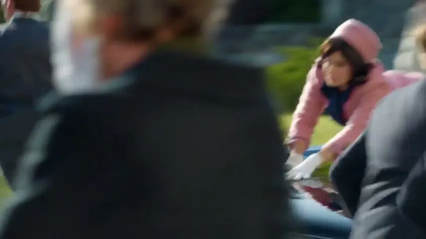 A woman in a pink suit and pillbox hat, resembling Jackie Kennedy, scrambles on the back of a moving car as men in suits surround her, in an exterior setting during the day.