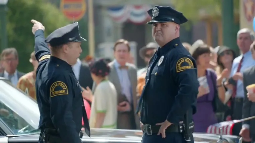 Two male police officers in uniform stand next to a car, looking up and talking to each other in a crowded street during the day.