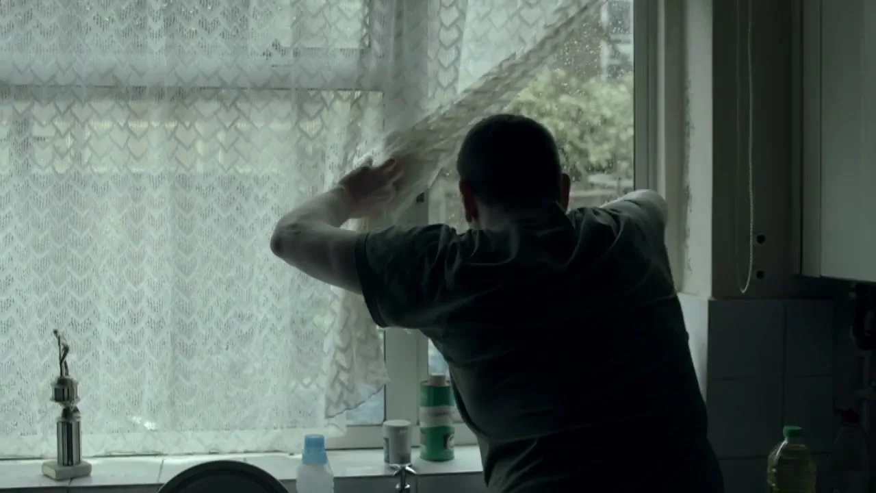 A man in a dark t-shirt looks out a window, pulling back a lace curtain in a dimly lit kitchen.