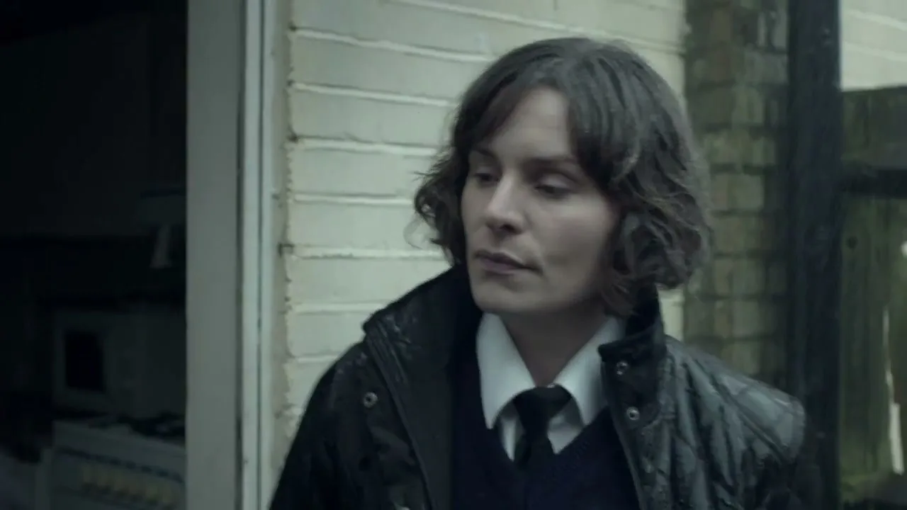A female animal control officer with short brown hair and a black jacket looks concerned while standing outside a house.