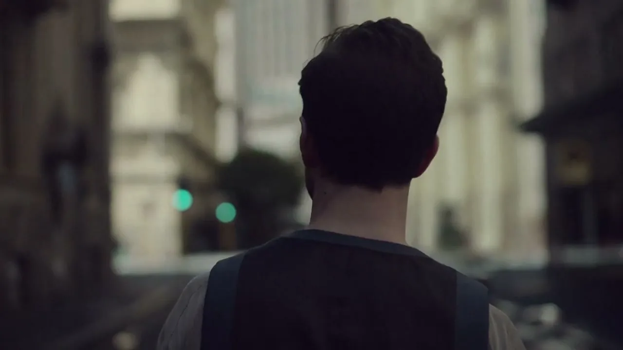 A man with dark hair, seen from behind, walks down a city street with tall buildings and a green traffic light in the distance.