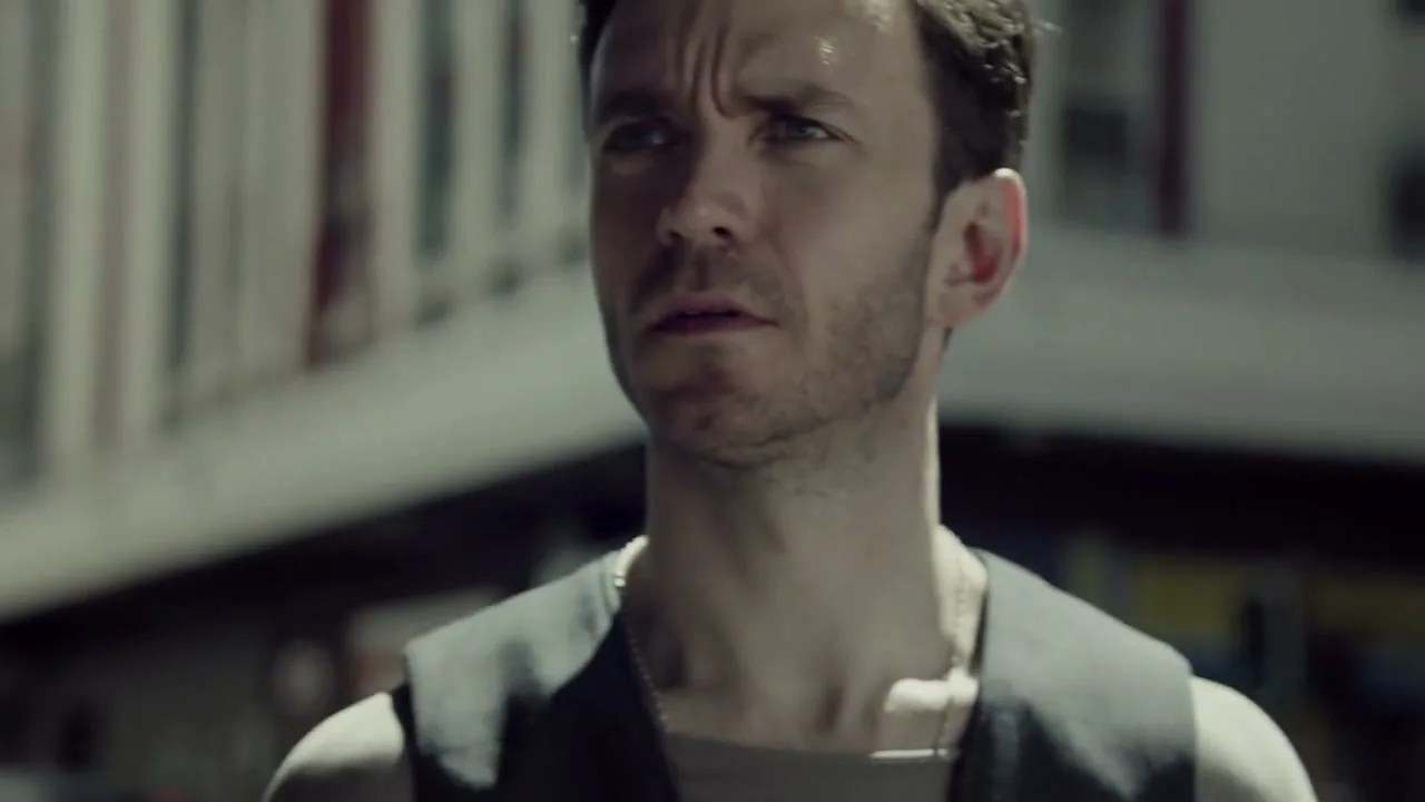 A man with short brown hair and a dark vest looks around with a concerned expression on a sunny street.