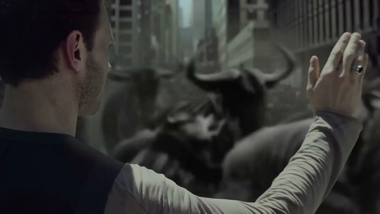 A man with a beard and a ring on his finger stands with his arm raised, facing a stampede of wildebeest in a city street.