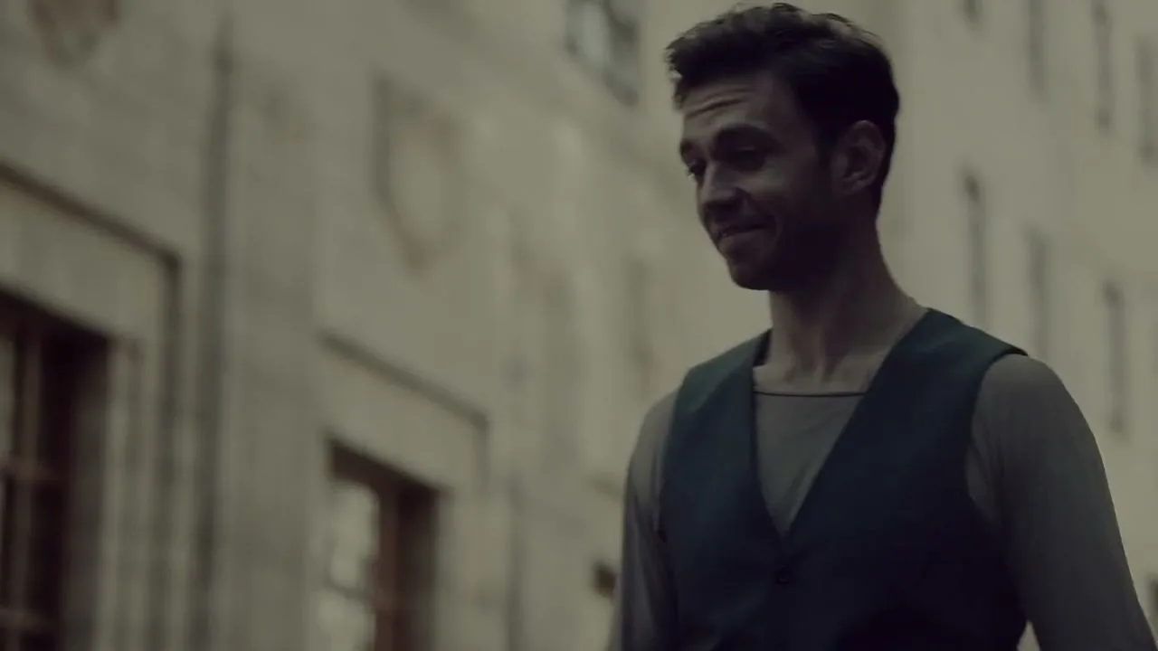 A man with dark hair and a dark vest smiles subtly, looking down and to the side, against a blurred building exterior.