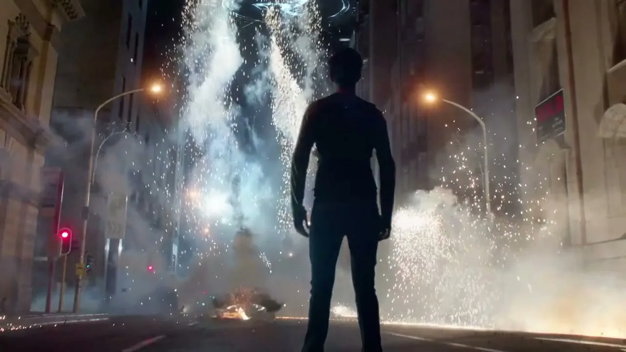 A man stands in the middle of a city street at night, looking up at a giant, glowing, alien-like structure amidst a shower of sparks and smoke.